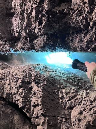 A flashlight illuminates a chunk of blue-tinted ice frozen in the permafrost layer, frozen dirt with grasses, rocks, and other material embedded in it.